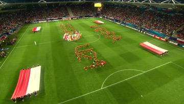 Ludzie uformowani w kształt orła i napisu "Polska" na zielonej murawie stadionu piłkarskiego, otoczeni przez kibiców na trybunach i flagi w narodowych barwach.