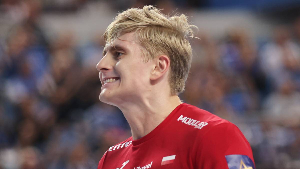 Mężczyzna w czerwonej koszulce sportowej z krótkimi blond włosami uśmiecha się podczas meczu piłki ręcznej.