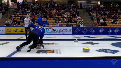 Gracz w curlingu z drużyny Szkocji zwalnia kamień na lodowisku, podczas gdy gracz z drużyny USA nadzoruje jego ruch. W tle widownia siedzi na trybunach.