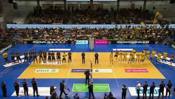 Dwie drużyny siatkarzy ustawione na boisku podczas meczu w wypełnionej po brzegi hali sportowej.