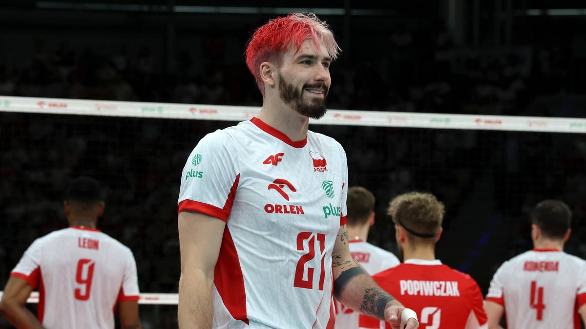 Siatkarz reprezentacji Polski w białej koszulce z czerwonymi elementami i numerem 21, z charakterystyczną fryzurą w czerwonym kolorze, uśmiecha się podczas meczu na tle innych zawodnik&oacute;w.