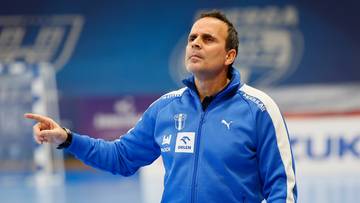 Trener w niebieskiej kurtce sportowej wskazuje palcem na hali sportowej.