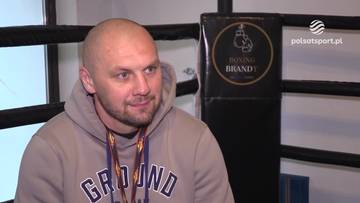 Portret Krzysztofa Głowackiego w szarej bluzie z kapturem, siedzącego na tle narożnika ringowego.