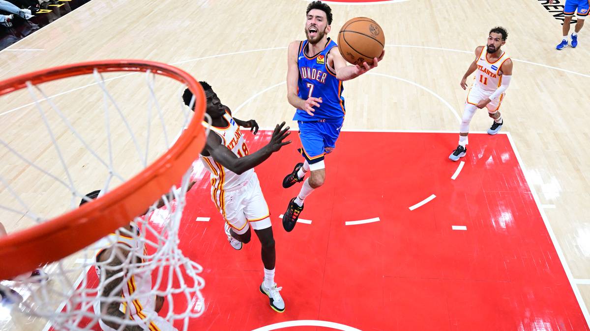 Zawodnik w niebieskim stroju z numerem 7 próbuje wykonać wsadu do kosza, podczas gdy zawodnik w białym stroju próbuje go zablokować. W tle widoczni są inni gracze.