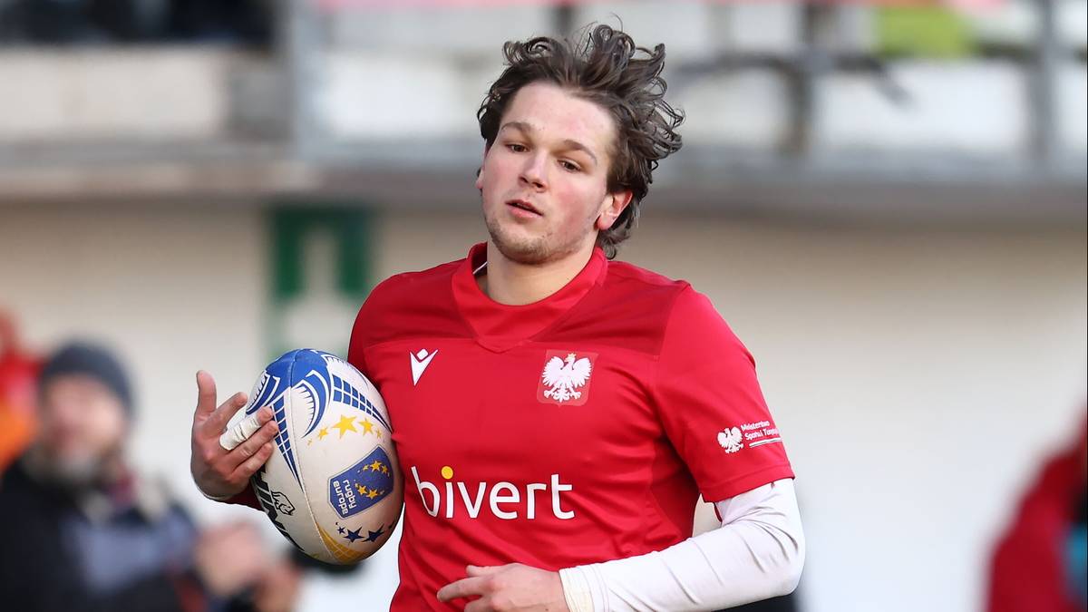 Młody zawodnik rugby w czerwonej koszulce z biało-czerwonym godłem i napisem "hivvert", trzymający piłkę do rugby.