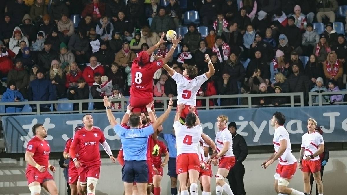 Gracze rugby wyskakują do piłki w powietrzu podczas meczu, z kibicami na trybunach w tle.