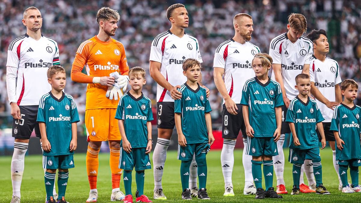 Grupa piłkarzy Legii Warszawa wraz z dziećmi w strojach sportowych, stojąca na murawie stadionu.