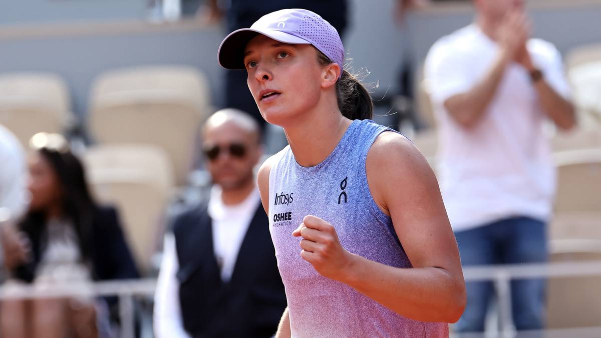 Iga Świątek, tenisistka, w okularach przeciwsłonecznych i czapce z daszkiem, z uniesioną ręką, ubrana w strój sportowy, na korcie tenisowym, podczas zawodów.