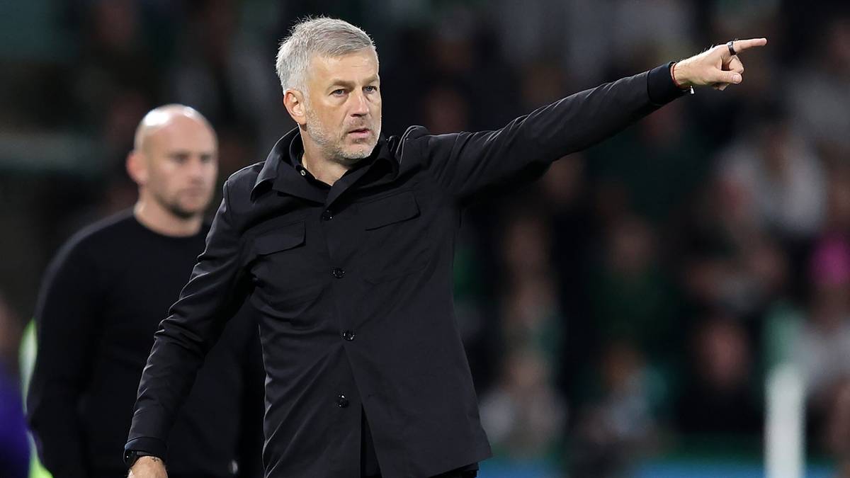 Trener piłkarski w czarnej koszulce ze wskazującym palcem skierowanym w bok, z tyłu inny mężczyzna.