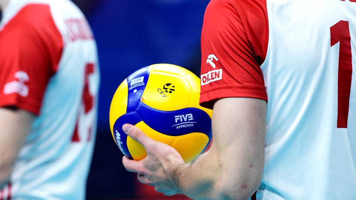 Gracz w białej koszulce z czerwonymi rękawami trzyma ż&oacute;łto-niebieską piłkę do siatk&oacute;wki.