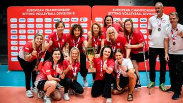 Grupa sportowc&oacute;w w czerwono-białych strojach pozuje z pucharem i medalami na tle czerwonej ścianki sponsorskiej po mistrzostwach w siatk&oacute;wce na siedząco.