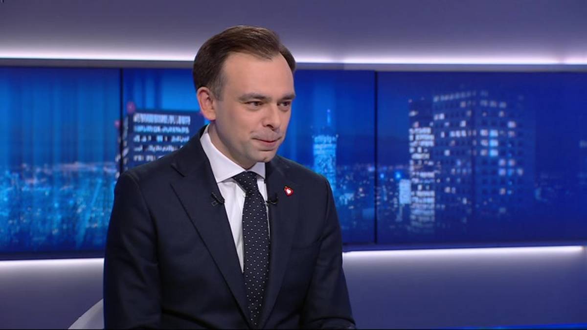 Mężczyzna w ciemnym garniturze i białej koszuli z krawatem siedzi i m&oacute;wi. Za nim widoczny jest ekran z nocnym widokiem miasta.