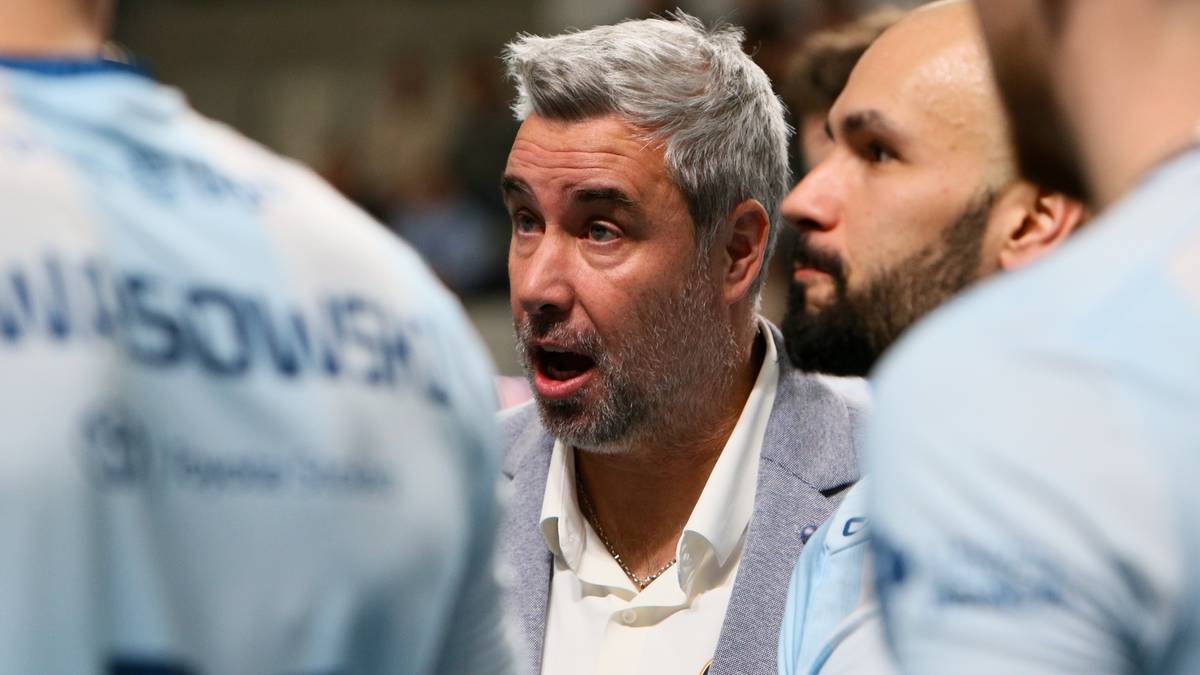 Trener siatkówki w średnim wieku z siwymi włosami i zarostem, w eleganckiej marynarce, przemawia do swoich zawodników.