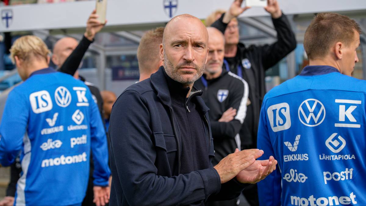 Mężczyzna w okularach przeciwsłonecznych i ciemnej kurtce, prawdopodobnie trener, stoi na tle innych mężczyzn w niebieskich koszulkach z białymi napisami.