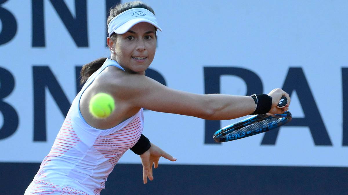 Tenisistka wykonuje uderzenie forehand, trzymając rakietę tenisową. Piłka tenisowa znajduje się w powietrzu obok jej ramienia. Kobieta ma na sobie białą koszulkę sportową i białą czapkę z daszkiem.