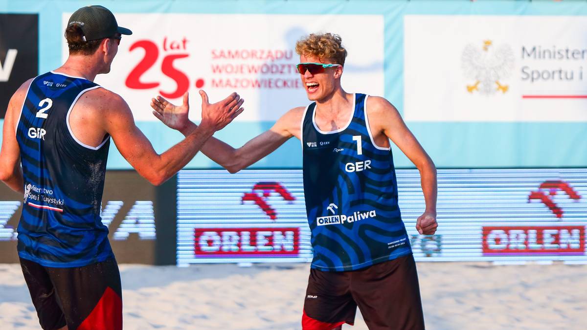 Dw&oacute;ch młodych mężczyzn w strojach do siatk&oacute;wki plażowej świętuje udaną akcję, podając sobie piątkę. Jeden z nich ma numer 7 na koszulce, drugi numer 2.