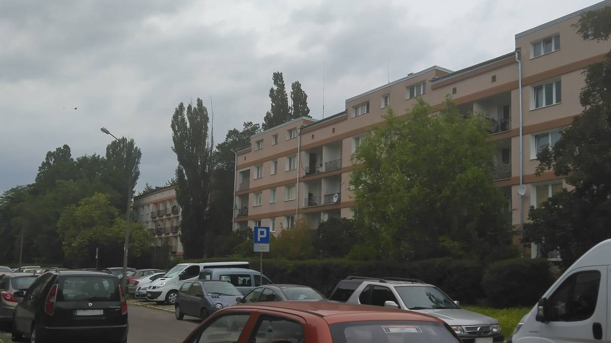 Widok ulicy z zaparkowanymi samochodami i blokiem mieszkalnym w tle.