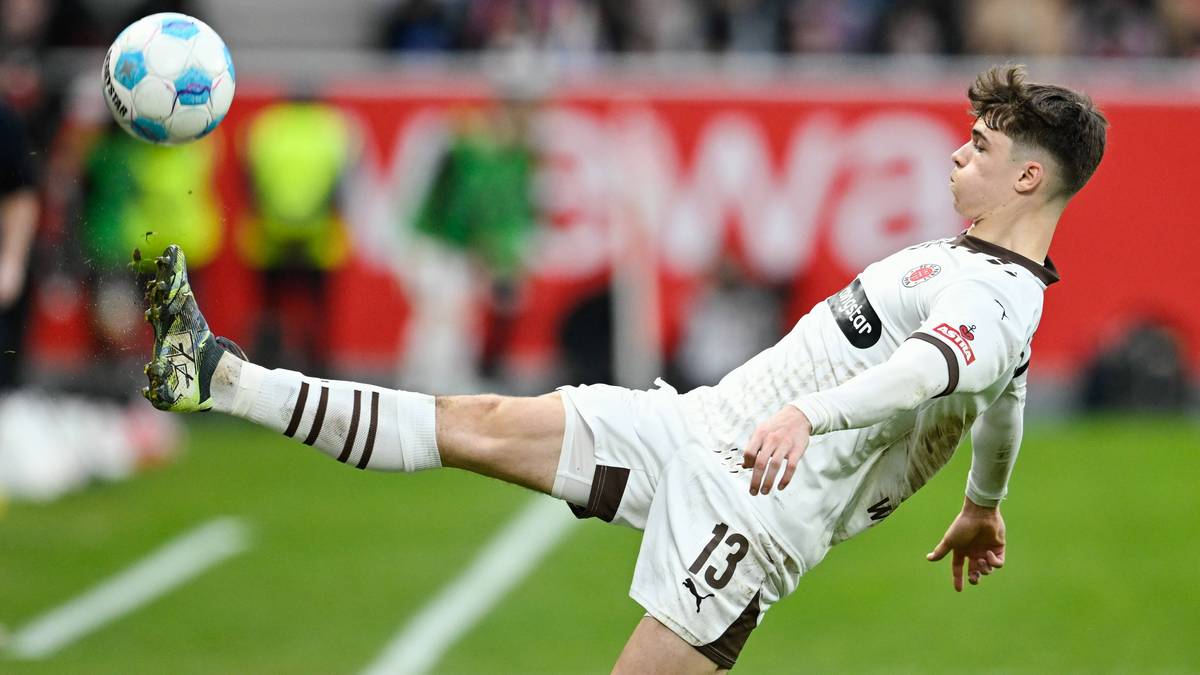 Młody piłkarz w białym stroju z numerem 13 wykonuje kopnięcie piłki nożnej podczas meczu. Piłka jest w powietrzu nad jego głową.