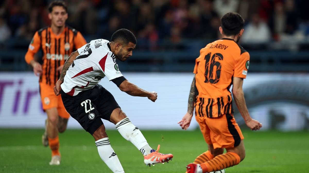 Piłkarz w białej koszulce z numerem 22 wykonuje strzał w kierunku bramki, a piłkarz w pomarańczowej koszulce z numerem 16 próbuje go zablokować podczas meczu piłki nożnej.