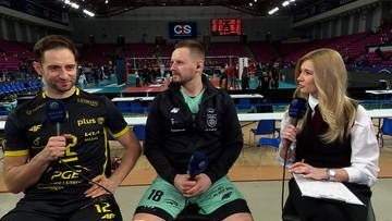 Dw&oacute;ch mężczyzn siedzi obok siebie, jeden trzyma mikrofon i uśmiecha się, drugi patrzy przed siebie. Obok nich siedzi kobieta prowadząca wywiad z mikrofonem i papierami.