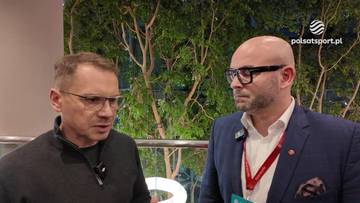 Dwóch mężczyzn w okularach rozmawia ze sobą. Jeden z nich ma na sobie czarny sweter, drugi granatowy garnitur z czerwoną wstążką na szyi.