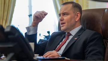 Mężczyzna w garniturze siedzi przy biurku i gestykuluje prawą ręką.