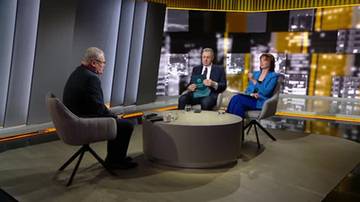 Trzy osoby siedzą przy okrągłym stole w studiu telewizyjnym. Dwóch mężczyzn i jedna kobieta biorą udział w dyskusji. W tle widoczne są nowoczesne ekrany.