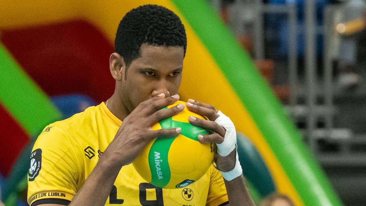 Siatkarz w ż&oacute;łtej koszulce z numerem 9 trzyma piłkę do siatk&oacute;wki przed twarzą w geście koncentracji.