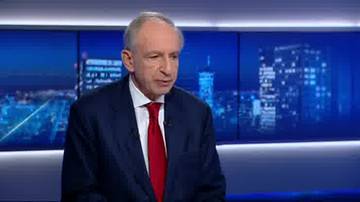 Starszy mężczyzna w ciemnym garniturze i czerwonym krawacie, wypowiadający się na tle nocnej panoramy miasta.