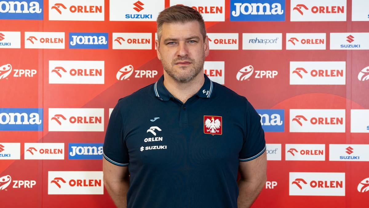 Mężczyzna w koszulce z godłem Polski i napisami sponsorskimi, stoi na tle ściany z logotypami sponsorów.