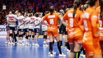 Drużyna piłki ręcznej kobiet w pomarańczowych strojach stoi na boisku, zwrócona plecami do widza.