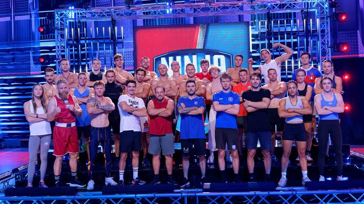 Grupa zawodników stoi na scenie przed wielkim ekranem z napisem "NINJA 10". Wszyscy ubrani są w sportowe stroje, niektórzy trzymają puchary i medale. Atmosfera jest uroczysta i radosna.