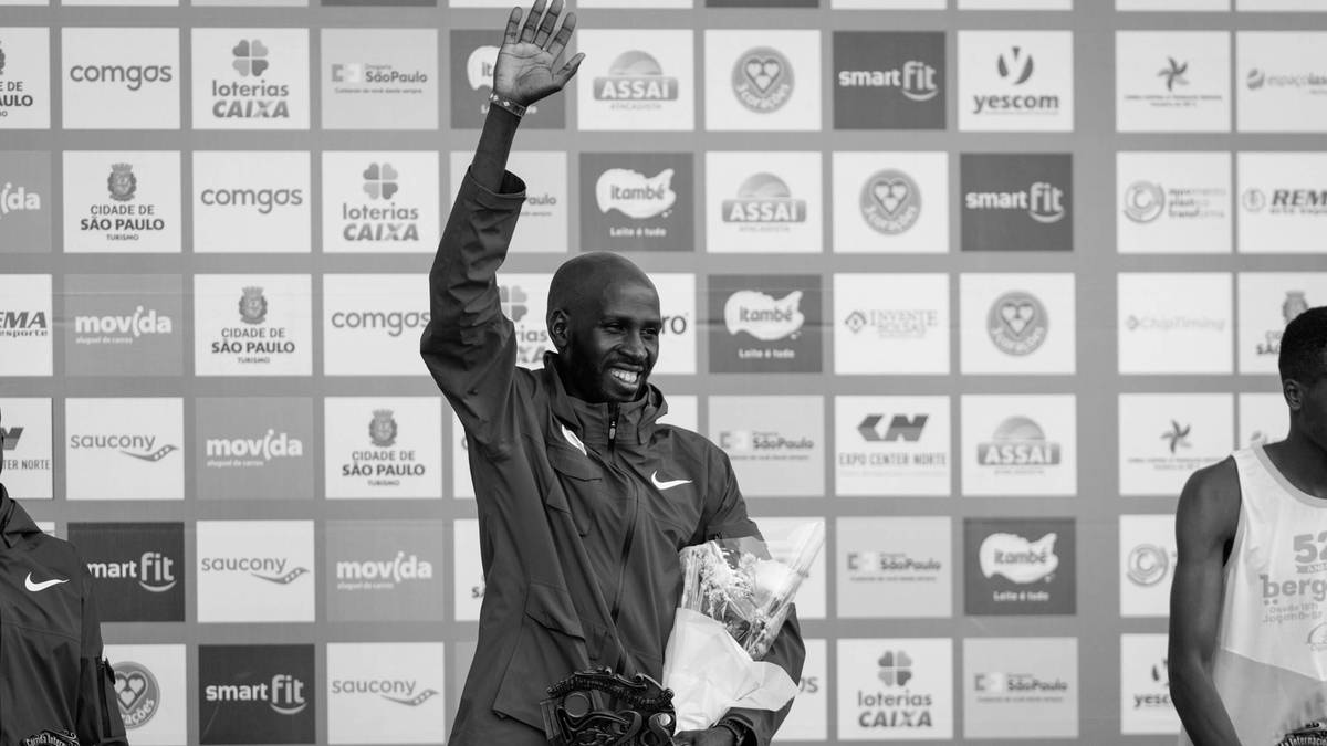 Mężczyzna w stroju sportowym, machający ręką, z bukietem kwiat&oacute;w w drugiej ręce, stoi na podium przed ścianą z logo sponsor&oacute;w.