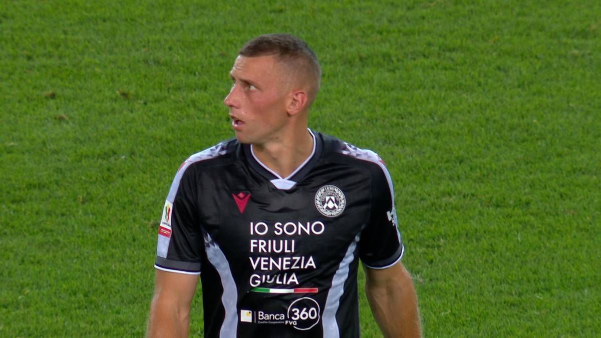 Piłkarz w czarno-białej koszulce z białym kołnierzykiem i rękawami, stoi na zielonej murawie stadionu piłkarskiego.