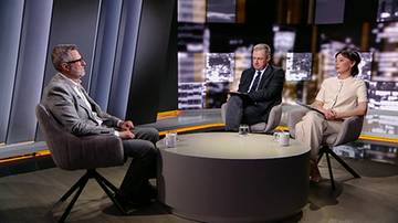 Trzy osoby siedzą w nowoczesnym studio telewizyjnym i rozmawiają przy okrągłym stole.