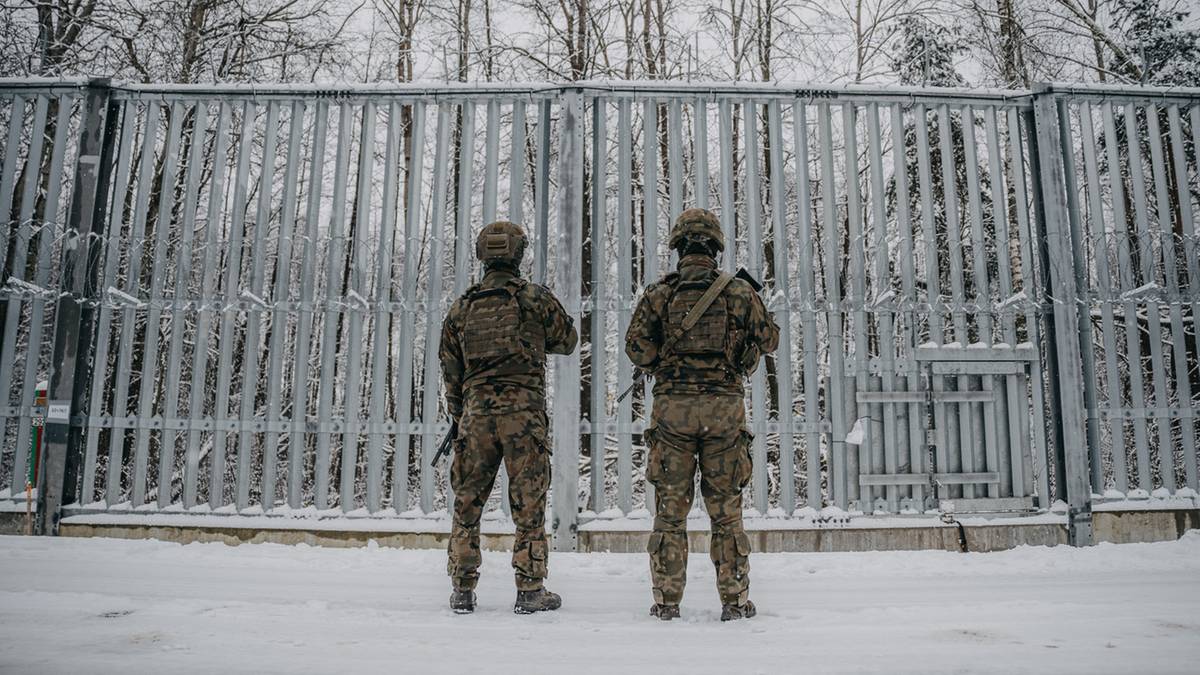 Dw&oacute;ch żołnierzy w mundurach stoi przed metalową bramą ogrodzenia.