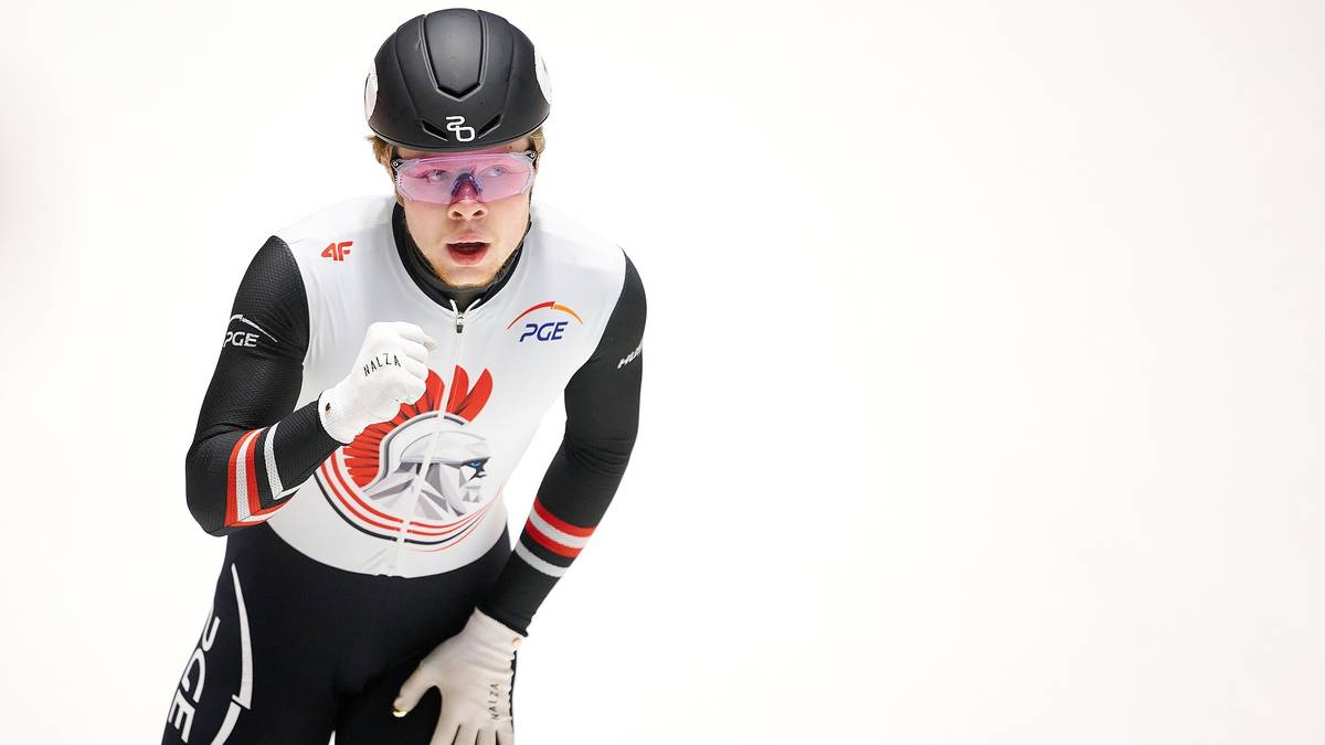 Łyżwiarz short track w czarno-białym stroju z czerwonym logo, z kaskiem i goglami, z wyrazem skupienia na twarzy, podczas biegu na lodzie.