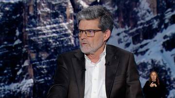 Mężczyzna w okularach i garniturze przemawia na tle g&oacute;rskiego krajobrazu.