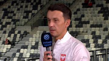 Mężczyzna w białej koszuli z polskim godłem trzyma mikrofon Polsat Sport na tle trybun stadionu.
