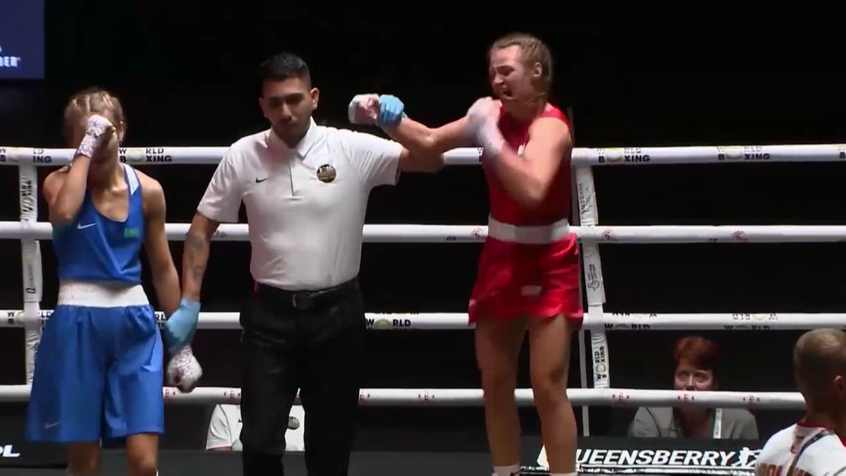 Julia Szeremeta w czerwonych spodenkach i rękawicach bokserskich unosi prawą rękę w geście triumfu. Obok niej stoi przegrana zawodniczka w niebieskim stroju, zasłaniająca twarz, oraz sędzia.