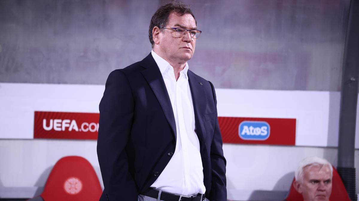 Mężczyzna w garniturze stoi na tle trybun stadionowych, z wyrazem skupienia na twarzy.