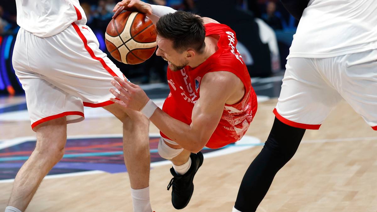 Koszykarz w czerwonej koszulce z piłką w rękach, próbujący obiec dwóch przeciwników w białych strojach.