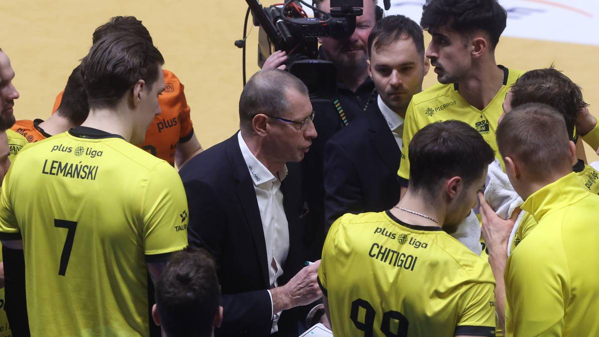 Grupa zawodników w żółtych koszulkach z numerami i nazwiskami, otaczająca trenera w garniturze, który pokazuje im coś na kartce.