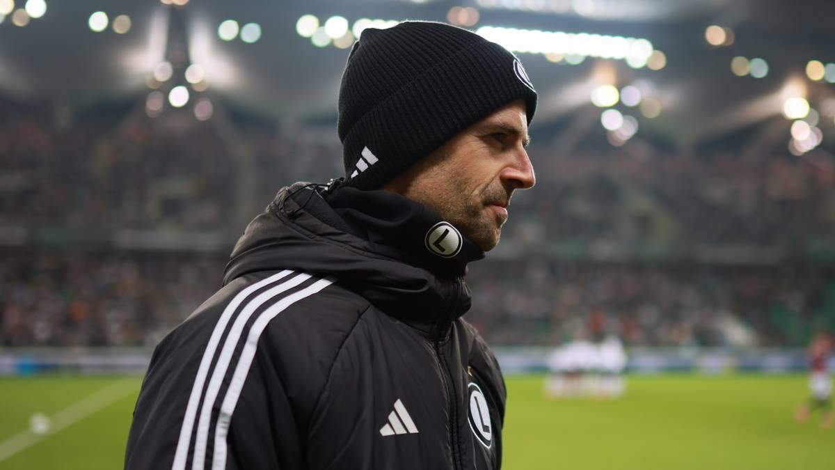 Trener piłkarski w czapce i czarnej kurtce z białymi pasami na ramionach stoi na tle stadionu piłkarskiego nocą.