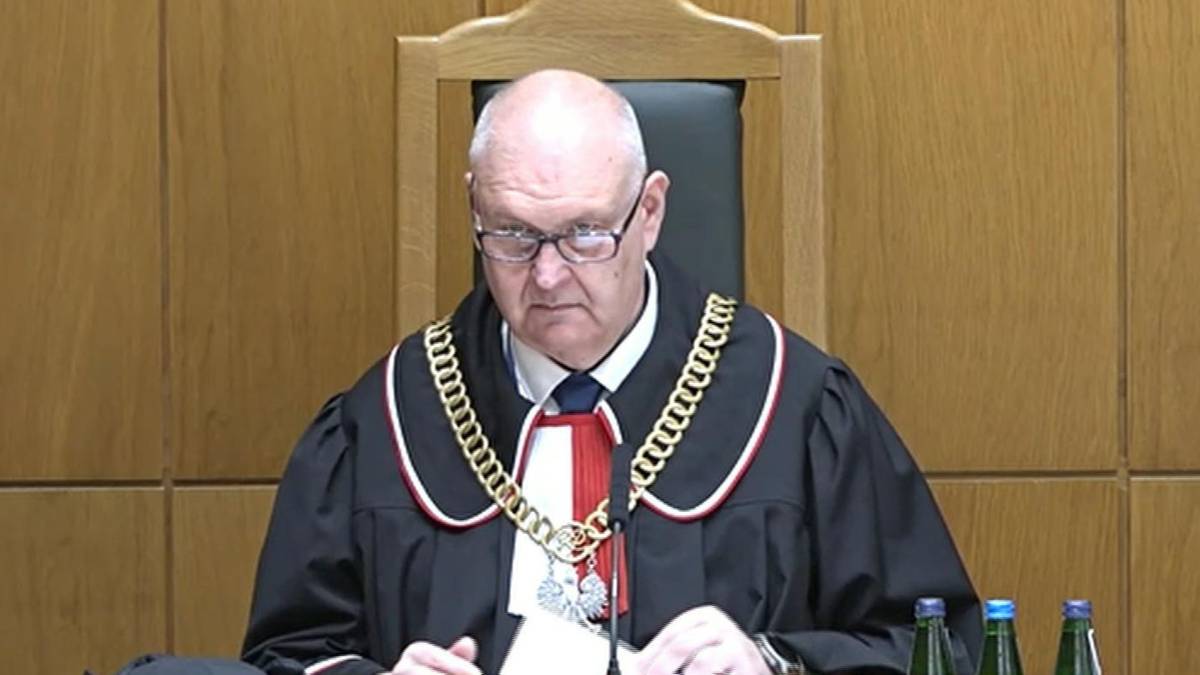 Sędzia w todze sądowej siedzi za pulpitem, przed nim mikrofon.