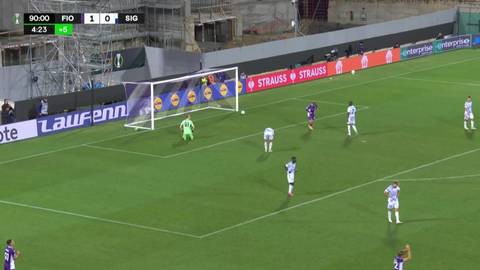 Gracze w białych i fioletowych strojach rywalizują na zielonej trawie podczas meczu piłki nożnej. Bramkarz w zielonym stroju broni bramki. Wynik na ekranie to 1:0 dla gospodarzy.