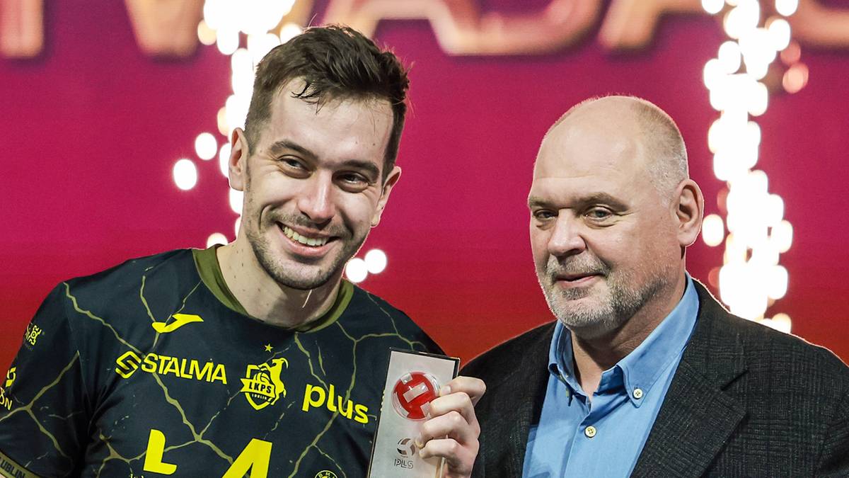 Dw&oacute;ch mężczyzn pozuje do zdjęcia. Młodszy mężczyzna po lewej stronie ma na sobie zielono-czarną koszulkę sportową z logo "STALMA PLUS". Trzyma w dłoniach białą kartkę z czerwonym symbolem. Starszy mężczyzna po prawej stronie ma na sobie ciemną marynarkę i niebieską koszulę. Obydwaj się uśmiechają.