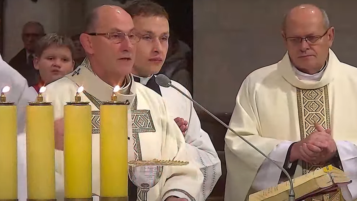 Ksiądz w szatach liturgicznych przemawia do mikrofonu, obok niego stoi drugi ksiądz. W tle widać uczestników nabożeństwa oraz cztery zapalone świece.