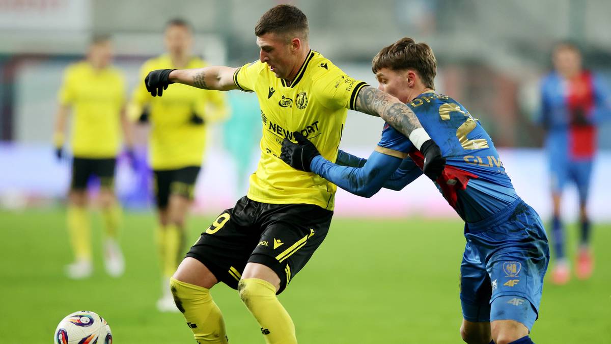 Piłkarz w żółtej koszulce walczy o piłkę z zawodnikiem w niebieskiej koszulce na boisku piłkarskim.