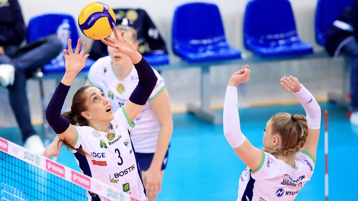 Dwie siatkarki w białych strojach sportowych podczas akcji meczowej, jedna próbuje zablokować piłkę, druga się do niej przygotowuje.